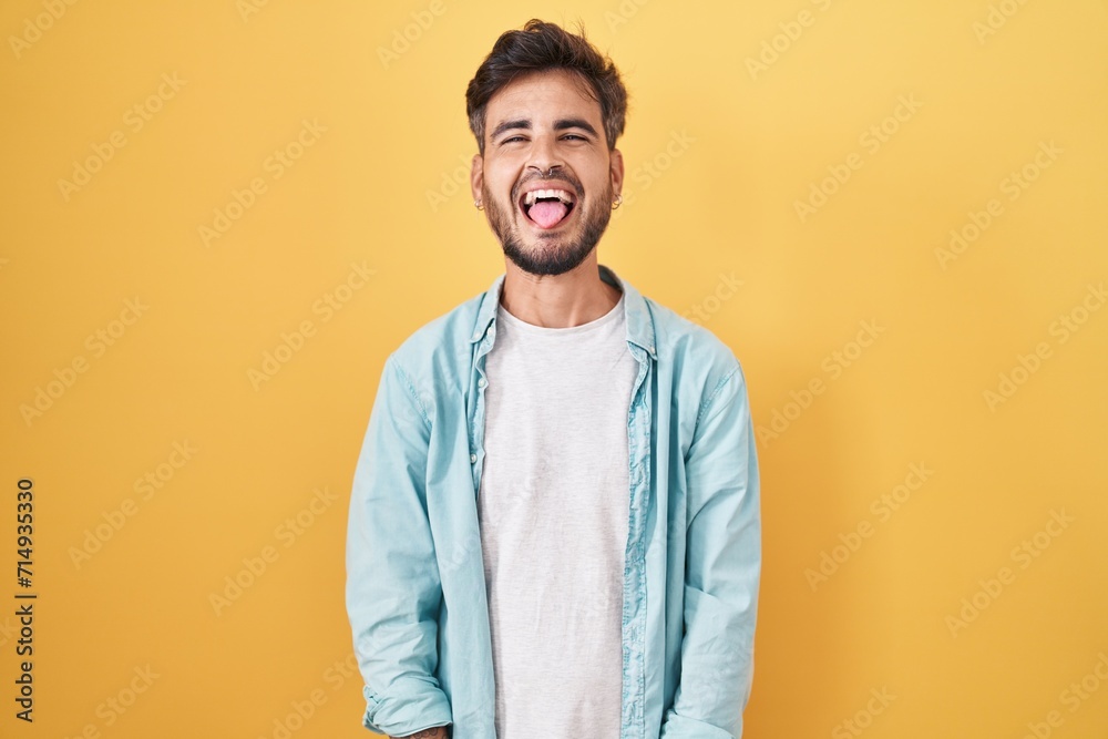Young hispanic man with tattoos standing over yellow background sticking tongue out happy with funny expression. emotion concept. - obrazy, fototapety, plakaty 