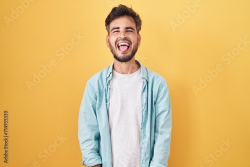Young hispanic man with tattoos standing over yellow background sticking tongue out happy with funny expression. emotion concept. photo