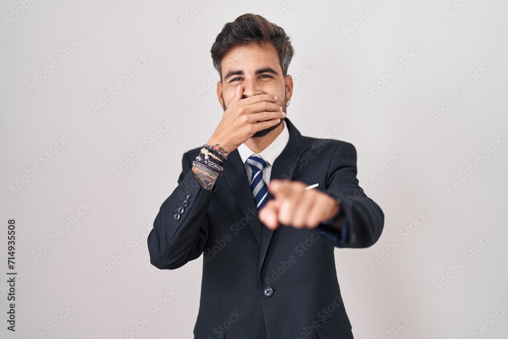 Young hispanic man with tattoos wearing business suit and tie laughing at you, pointing finger to the camera with hand over mouth, shame expression Young hispanic man with tattoos wearing business suit and tie laughing at you, pointing finger to the camera with hand over mouth, shame expression