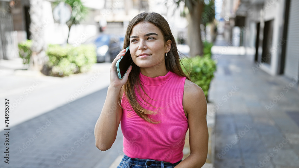 Basking in sunlight, a young and beautiful hispanic woman exudes confidence, her joyful smile radiates happiness as she casually engages in a fun, lively street-side chat on her smartphone.