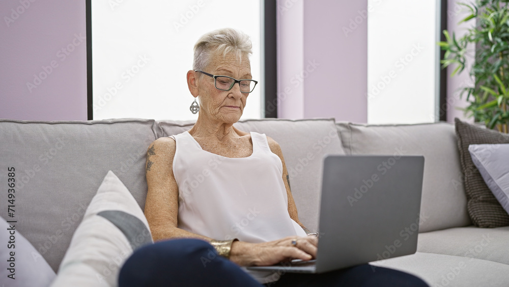 Focused elderly woman with grey hair and glasses, using laptop with concentration while sitting on sofa indoors at home, embodying mature technology use in the living room Focused elderly woman with grey hair and glasses, using laptop with concentration while sitting on sofa indoors at home, embodying mature technology use in the living room