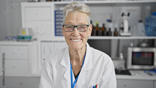 Confident and smiling grey-haired senior woman scientist enjoying her work  sitting happily at the lab amidst microscope and test tubes