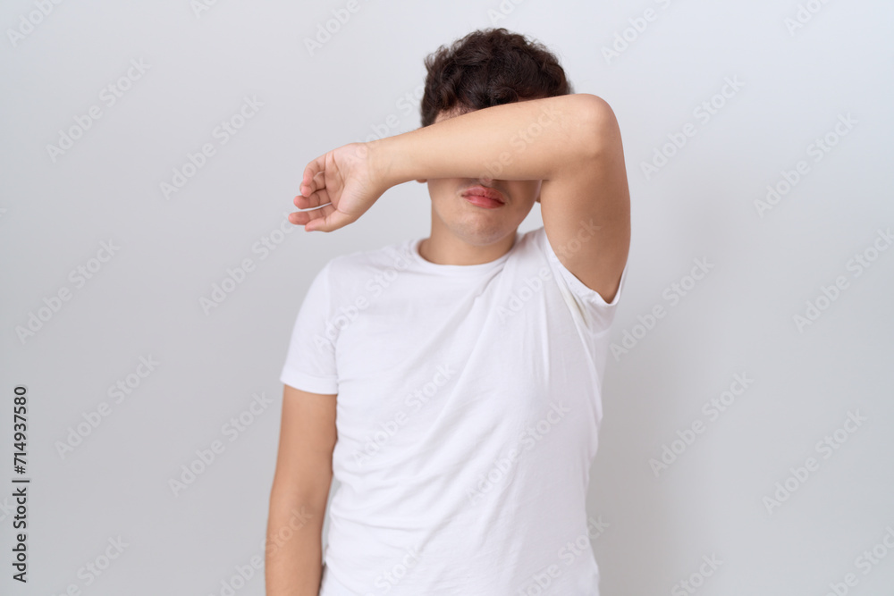Young non binary man wearing casual white t shirt covering eyes with arm, looking serious and sad. sightless, hiding and rejection concept