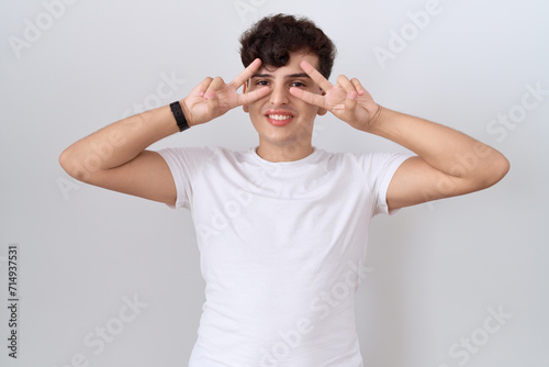Young non binary man wearing casual white t shirt doing peace symbol with fingers over face, smiling cheerful showing victory © Krakenimages.com