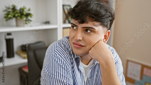 A young, thoughtful man in a striped shirt daydreaming in a modern office, portraying casual professionalism and a relaxed work environment.
