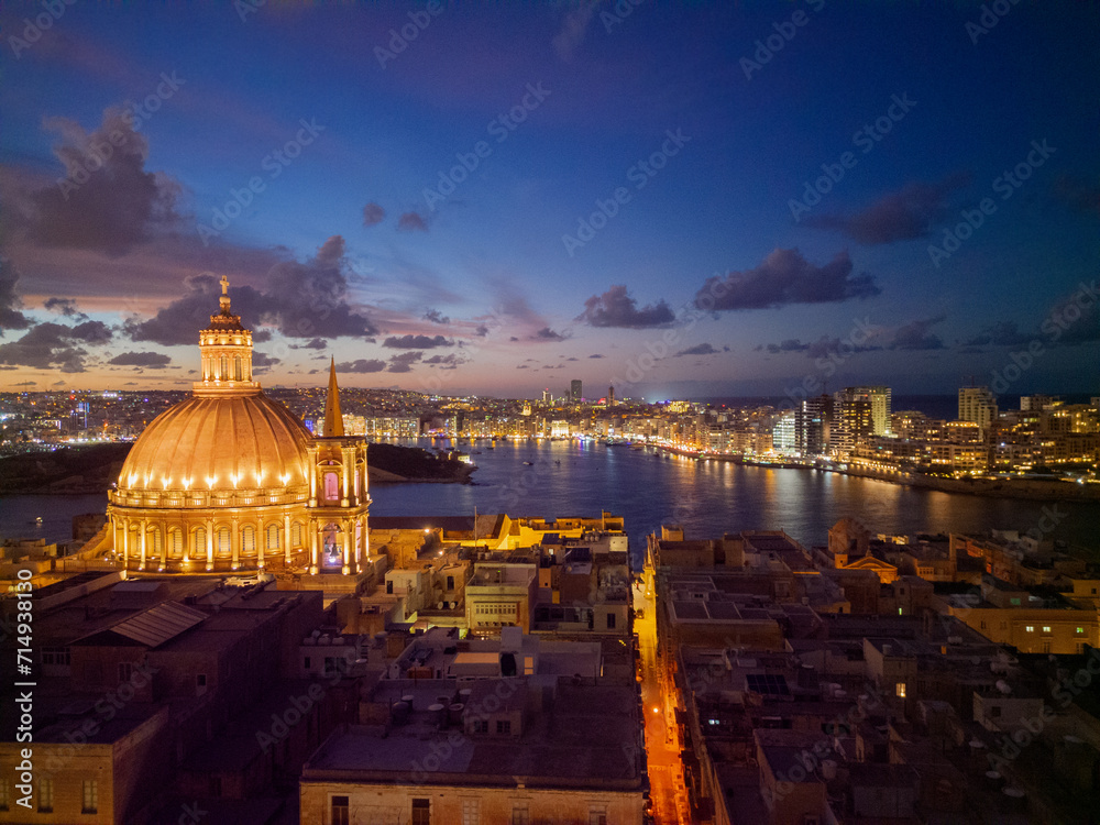 Beautiful illuminated dome of Basilica of Our Lady of Mount Carmel in Valletta. Aerial view with bright orange street line towards Sliema skyline and light reflection in the sea.