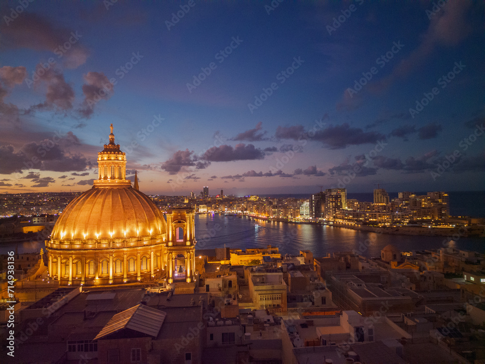 Illuminated golden color dome of Basilica of Our Lady of Mount Carmel at dawn. Aerial view form historic Valletta town towards Sliema skyline with modern buildings and light reflection on the sea.