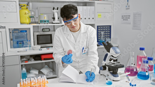 A young man examines a petri dish in a laboratory filled with scientific equipment, portraying a professional medical or research setting.