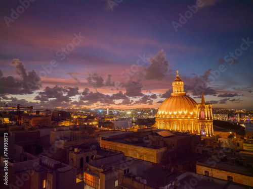 Sunset with pink sky in Valletta. Basilica of Our Lady of Mount Carmel in golden color with illuminated dome. Beautiful bright orange curtain window from a residential apartment.