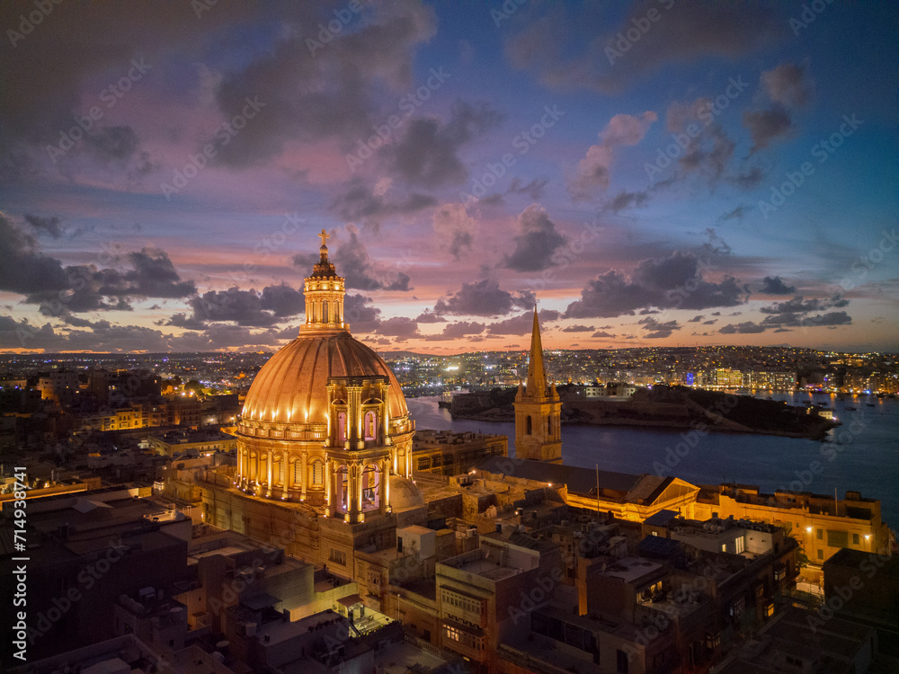 Illuminated dome of Basilica in historic Valletta town at sunset with pink sky. Aerial view towards Manoel island in the dark.