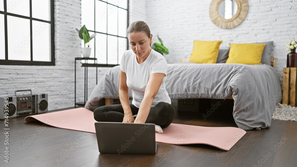 Captivating young blonde woman enjoying a morning yoga class online, lying on a cozy bed with her laptop on the bedroom floor
