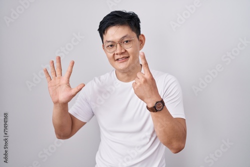 Young asian man standing over white background showing and pointing up with fingers number six while smiling confident and happy.