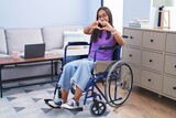Young hispanic woman sitting on wheelchair at home smiling in love doing heart symbol shape with hands. romantic concept.