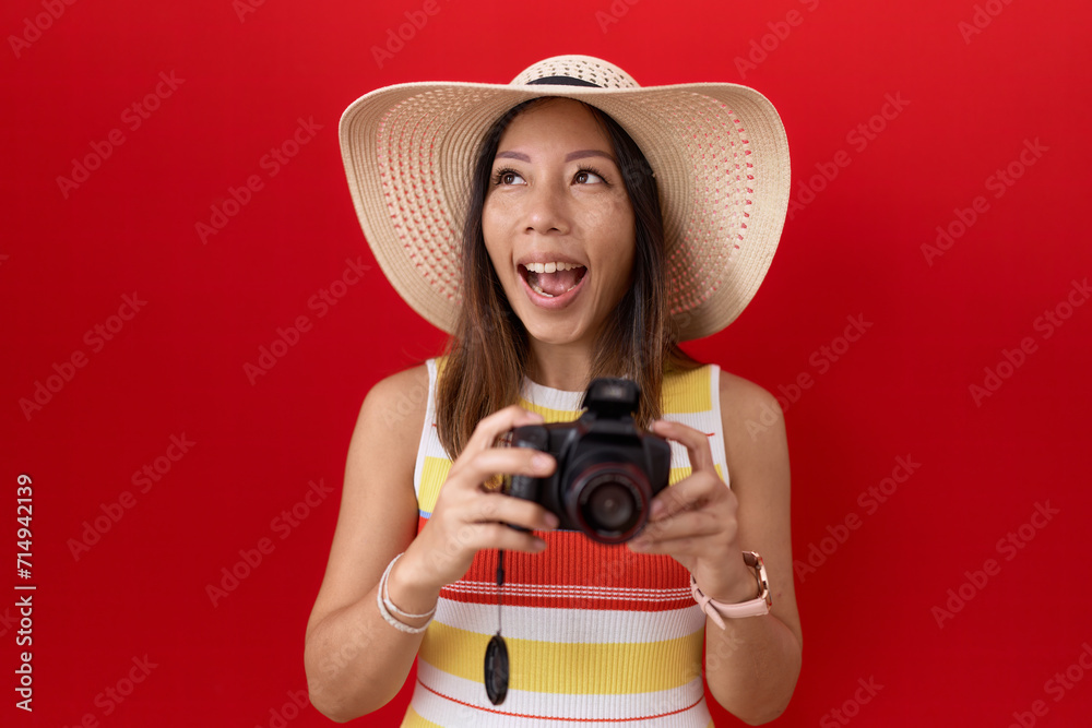 Middle age chinese woman using reflex camera wearing summer hat angry and mad screaming frustrated and furious, shouting with anger looking up.