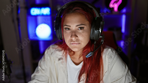 Focused young redhead woman streamer, engrossed in intense gaming session, in a serious virtual face-off, headset on, encapsulated in the dimly-lit indoors of her home gaming room photo