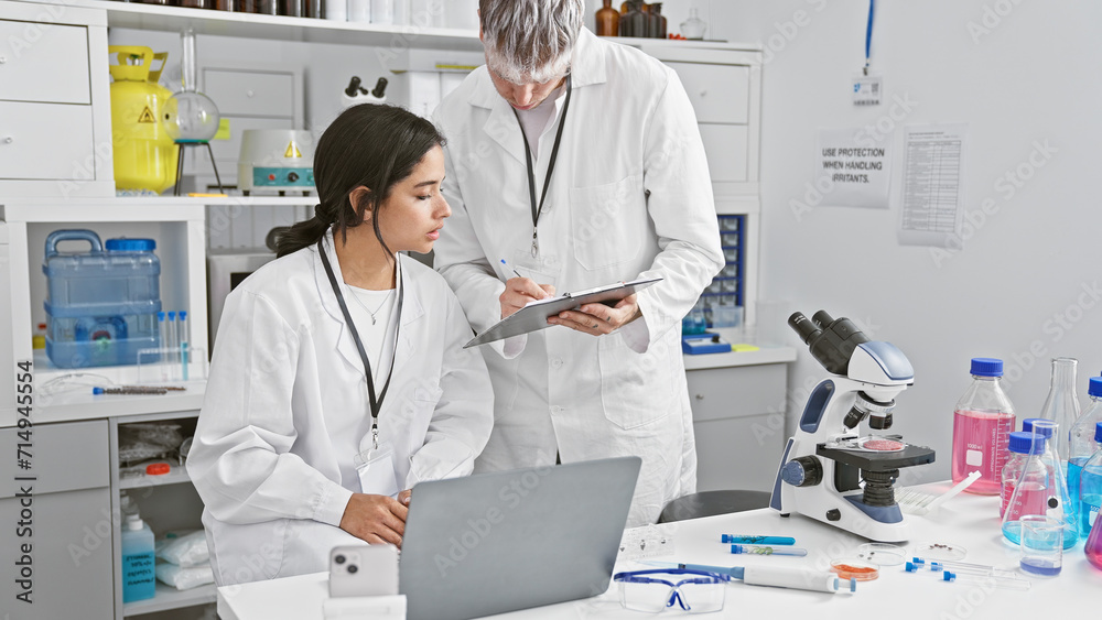 A woman and man, seemingly coworkers, analyze data together on a laptop in a well-equipped laboratory setting.