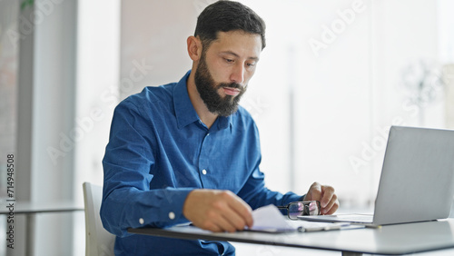 Young hispanic man business worker using laptop reading document at the office