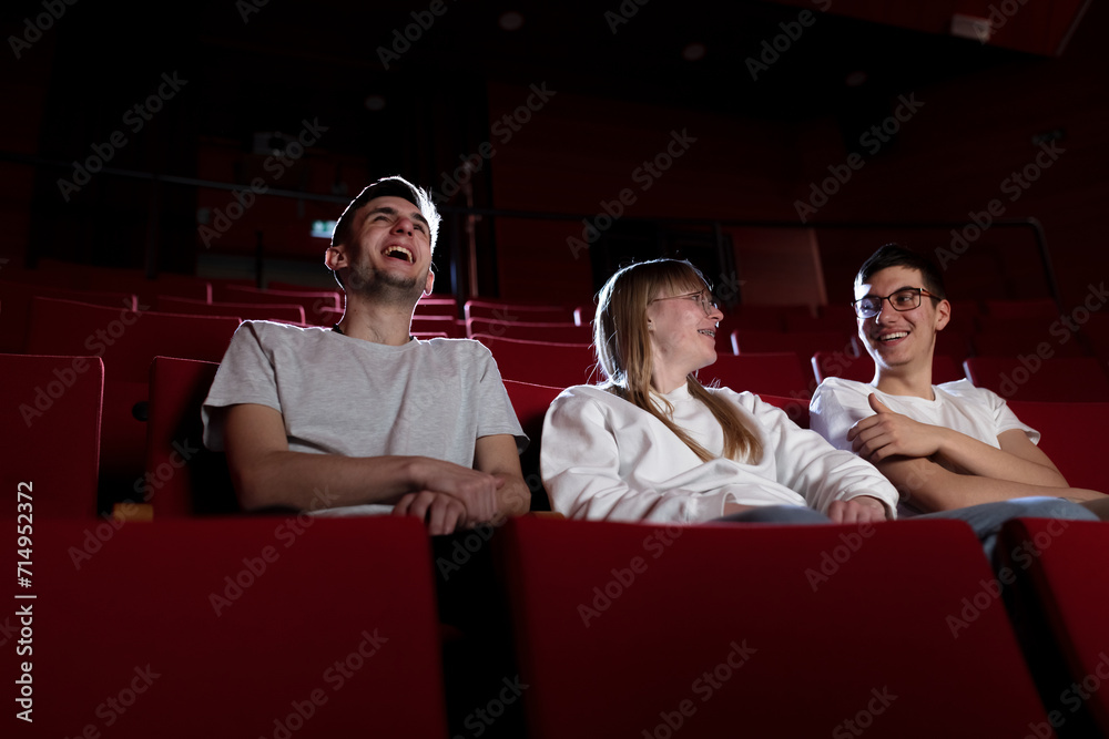 Three friends, a teenage girl and two boys smiling, watching a movie in an empty cinema with red chairs. Young adults and leisure concepts. - obrazy, fototapety, plakaty 