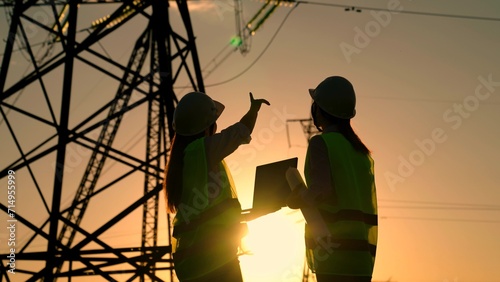 Teamwork of power engineers with computer in protective helmets, maintenance of power lines in outdoors. Two Female civil engineers work together using computer on power line. Green energy. Builders