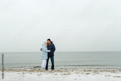 An elderly couple stands by the seashore in winter, hugging.