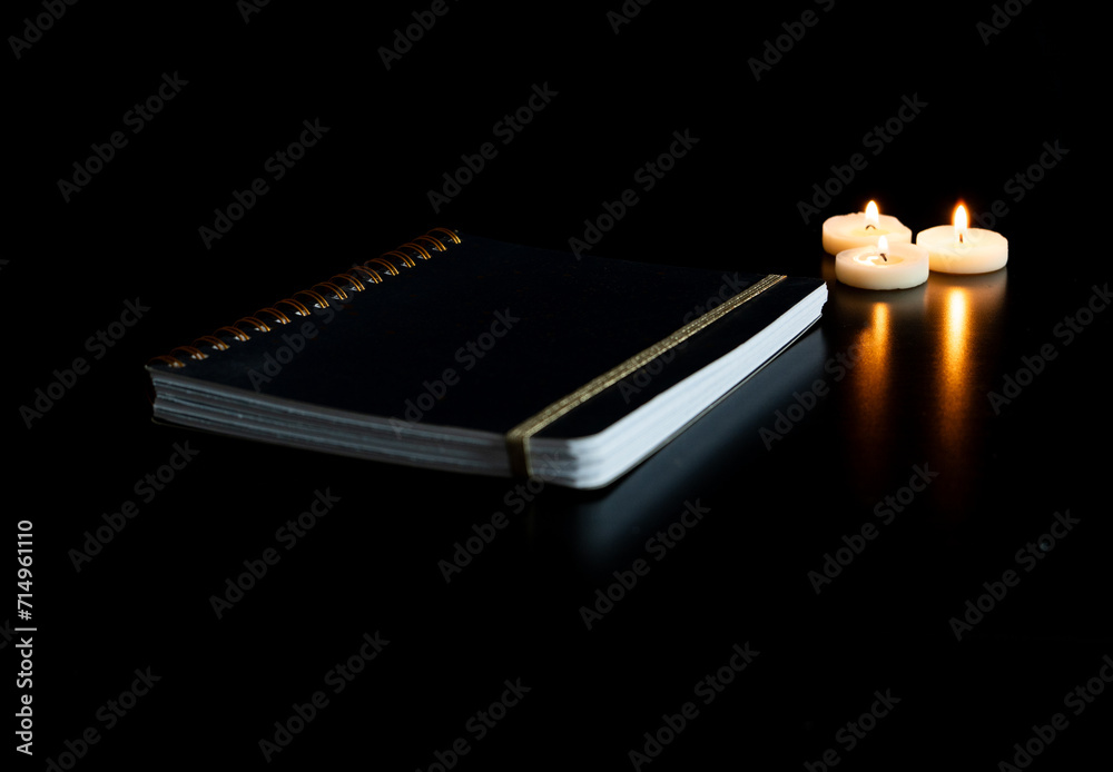 Cahier fermé sur une table noire avec 3 petites bougies chauffe plat allumées et leur reflet - fond noir et espace vide Cahier fermé sur une table noire avec 3 petites bougies chauffe plat allumées et leur reflet - fond noir et espace vide