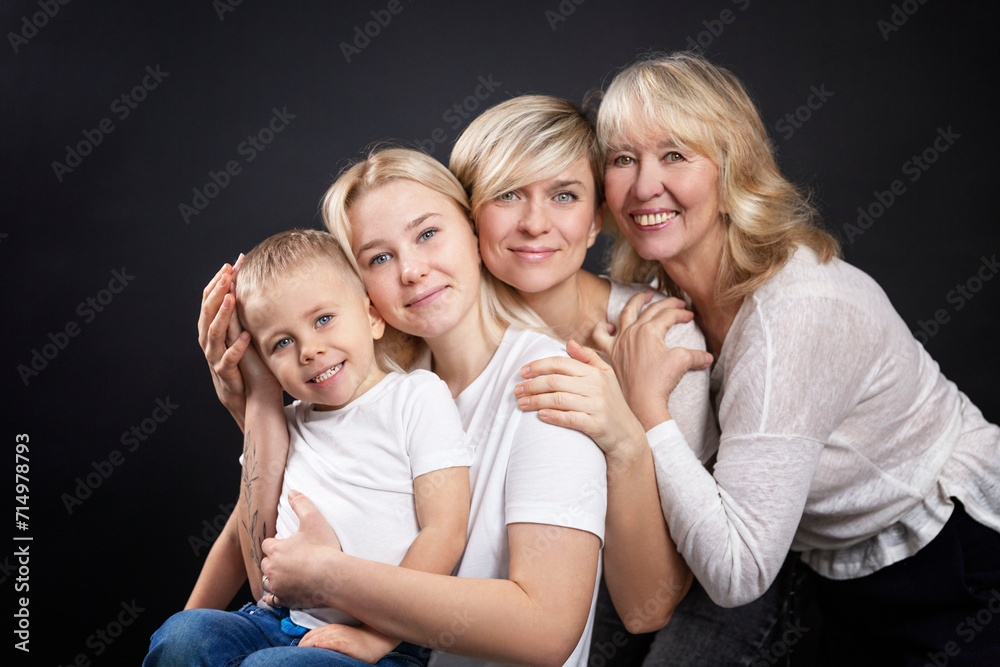 Portrait of three generations of women and a little boy. Grandmother, mother, daughter and son in white T-shirts. Love and tenderness. Black background. Portrait of three generations of women and a little boy. Grandmother, mother, daughter and son in white T-shirts. Love and tenderness. Black background.