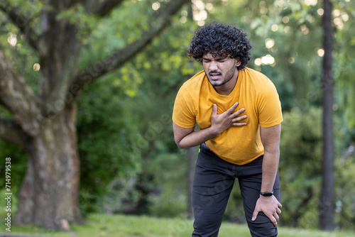 Tired young Indian male athlete standing bent over in park and holding hand to chest, feeling severe pain and shortness of breath after jogging photo