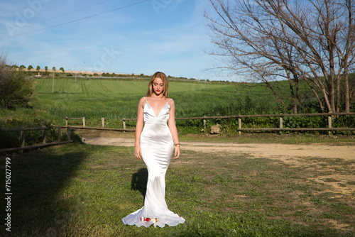 A beautiful young woman, blonde and with long hair, wearing a white dress and a crown of flowers in her hair, is in a field. In the background a green wheat field. The woman is in different poses.