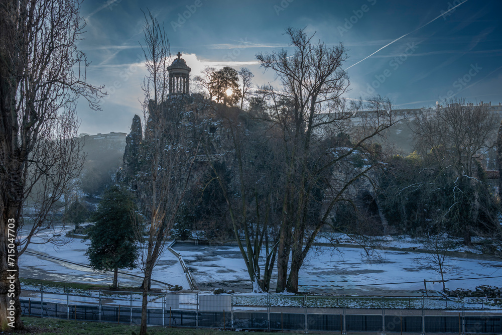 Paris, France - 01 20 2024: Park des Buttes Chaumont. View the central part of the park with footbridge, belvedere island, Temple of the Sibyl and the lake under under the snow.