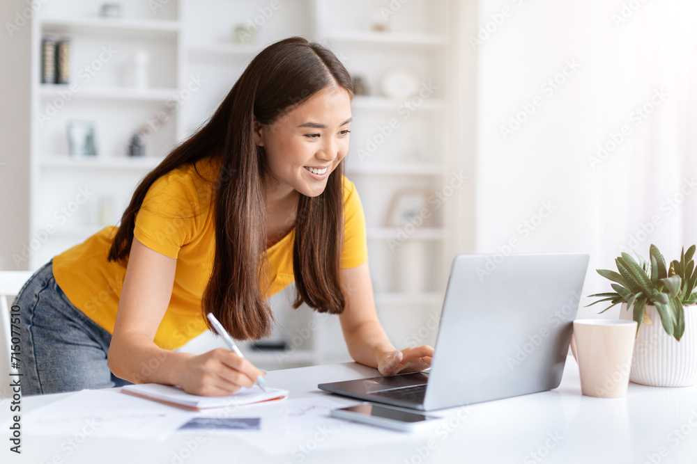 Focused Asian woman working on laptop and taking notes at home office Focused Asian woman working on laptop and taking notes at home office