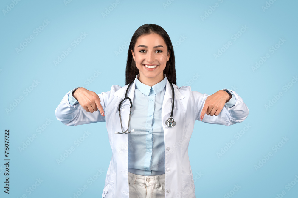 Friendly european female doctor in white coat pointing down at free space and smiling at camera on blue background Friendly european female doctor in white coat pointing down at free space and smiling at camera on blue background