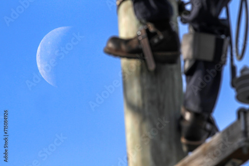 Photo of a lineman climbing a pole in boots and long pants during daylight while the moon rises. 