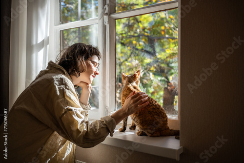 Breed cat Devon Rex sitting on windowsill, warming in sunlight. Loving teenager looking at kitty with love, tenderness. Teen caressing, stroking kitten purring of pleasure. Unconditional love to pets
