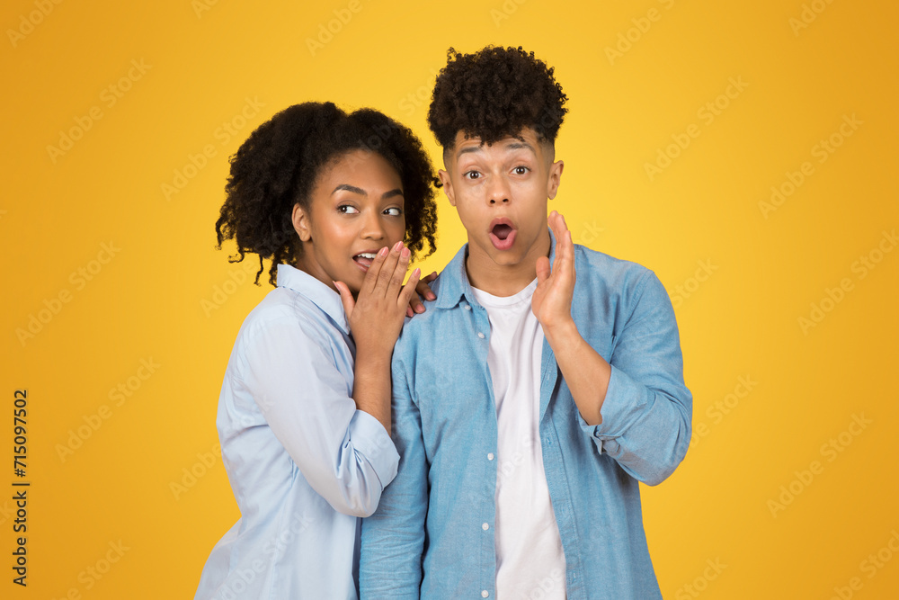 A young woman whispers into her hand with a mischievous expression while a young man