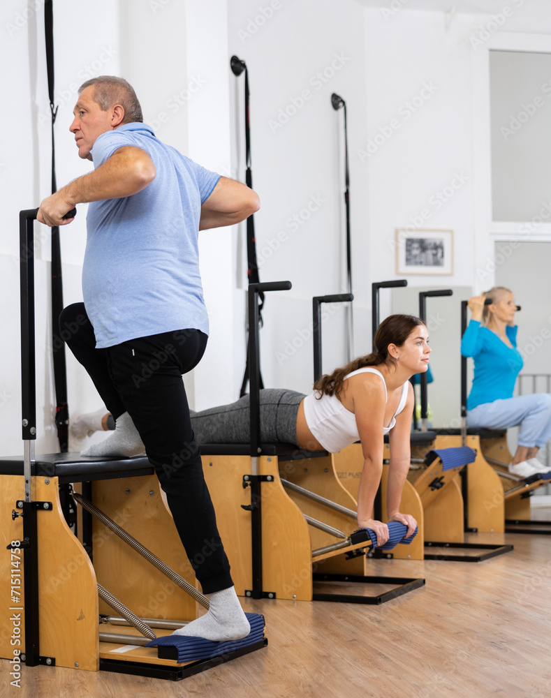 Fototapeta premium Serious concentrated elderly man wearing sportswear doing standing exercises on Pilates chair machine in rehabilitation studio