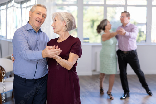 Happy smiling elderly woman enjoying impassioned merengue with male partner in modern latin dance salon. Social dancing concept