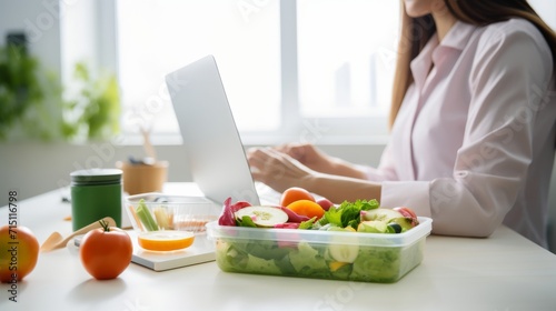 Female office worker at her desk  with healthy  lunch of fruit and vegetables in a Tupperware container corner of desk. Theme healthy  eating