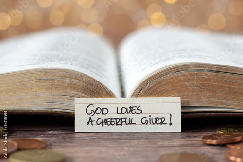 God loves a cheerful giver, handwritten text note in front of open holy bible book and golden coin money with bokeh background. Christian biblical concept of giving, tithing, and religious offering.