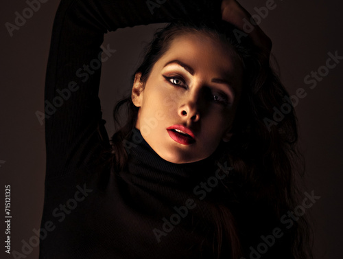 Beautiful makeup woman with red lipstick in black poloneck posing on dark shadow studio black background. Long brown hairstyle. Closeup photo