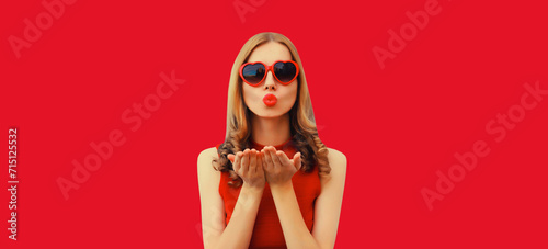 Portrait of beautiful young woman blowing her lips with red lipstick sends sweet air kiss wearing heart shaped sunglasses on studio background