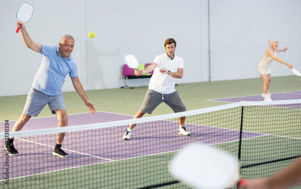 Obraz premium Portrait of sporty senior man playing doubles pickleball with male partner on indoor court, ready to hit ball. Sport and active lifestyle concept..