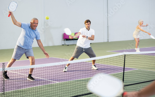 Portrait of sporty senior man playing doubles pickleball with male partner on indoor court, ready to hit ball. Sport and active lifestyle concept..