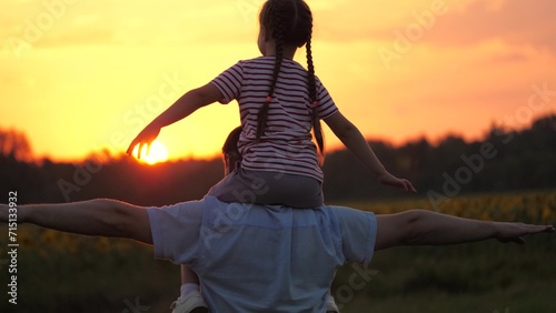 Father with daughter silhouette on shoulders pretends to be plane extending arm. Father lifts arms mimicking flight of airplane with girl enjoying shoulder ride. Father carries daughter on shoulders