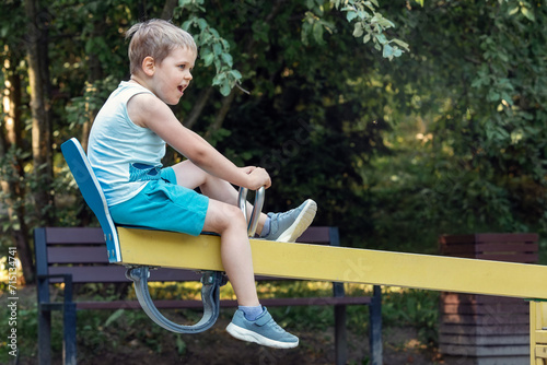 Little boy on yellow balance seesaw in green park foliage background