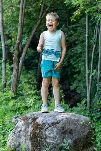 A small boy climbs on a big stone in the forest and shouts loudly