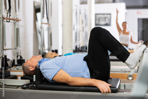 Focused mature man lying with her legs and arms suspended while doing Pilates exercise on reformer bed equipment in studio