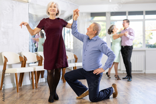 Happy mature couple enjoying retro ballroom dancing in modern dance salon, man standing on knee and holding hand of woman photo