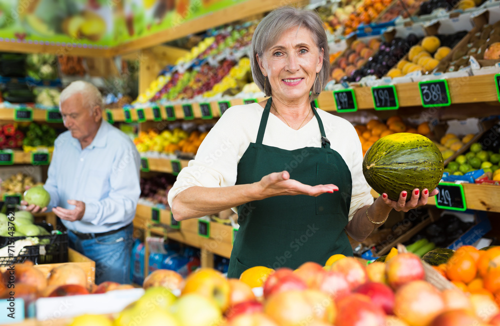 Mature woman greengrocer worker standing amongst shelves with fruits and vegetables and holding melon in hands. Old man making purchases in background.
