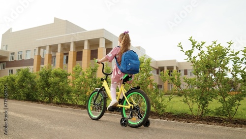 Little girl pupil with backpack riding to elementary school on bike at schoolyard road with trees slowmo. Active female kid child driving on cycle transportation arriving to classroom campus