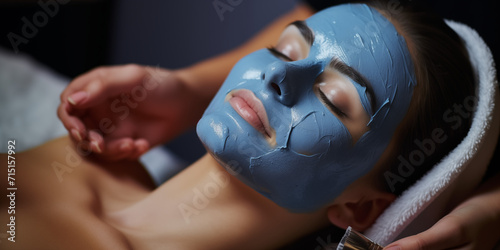 A young woman at a beauticians appointment. The specialist applies a cream mask to the face. Facial skin care and protection. A girl at the cosmetologist's office. Face in blue clay. Care procedure. B